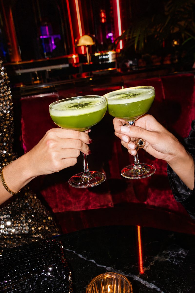 two friends clinking green matcha cocktails at a moody bar